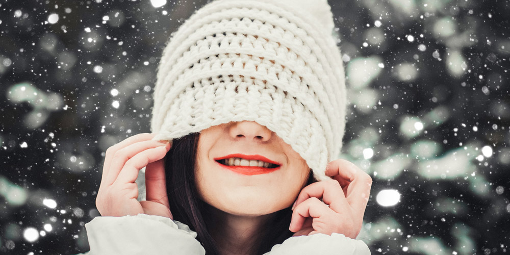 Woman outside in snow pulls down white knit hat over her eyes and smiles