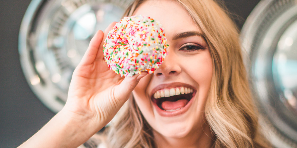 Woman with long, blond, wavy hair smiles at the camera and holds donut with rainbow sprinkles in front of one eye