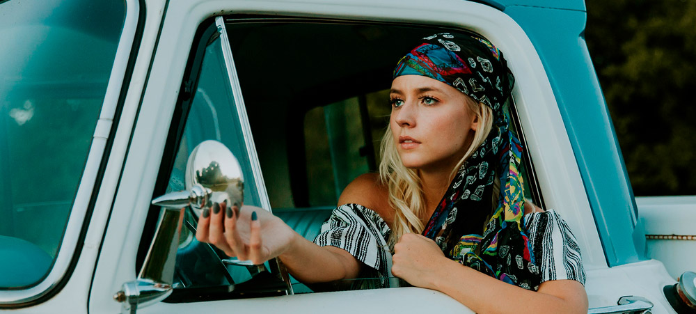 Woman sits in vintage blue pickup truck adjusting side mirror through the open window; a colorful scarf is tied around her long, blonde hair