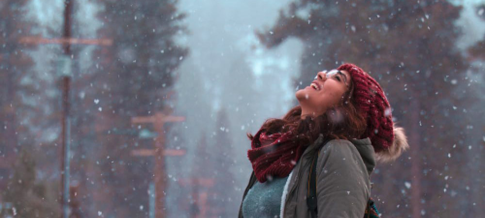 woman stands outside while it’s snowing and looks up to the sky, lauging