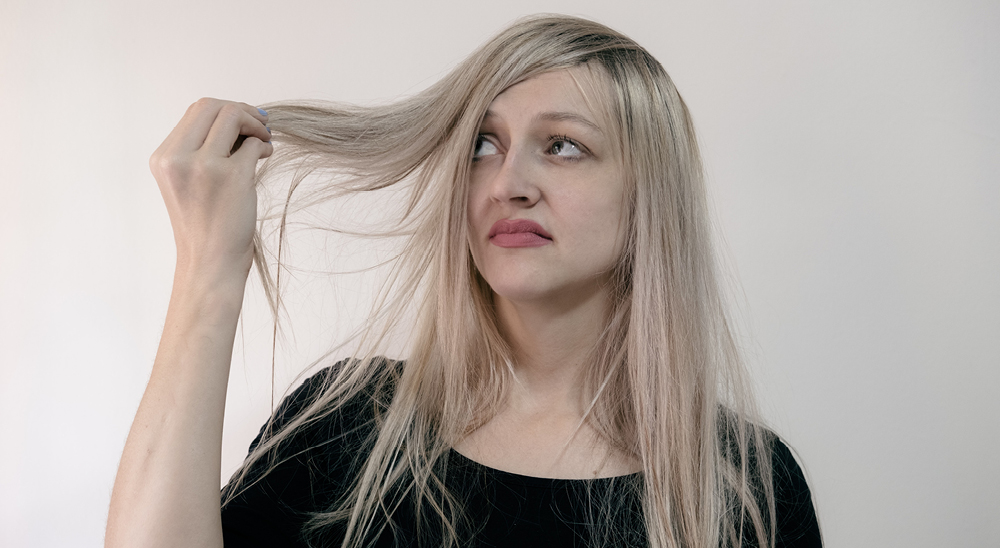 woman with light blonde hair against gray backround holds her stringy, dry hair with a look of disgust on her face